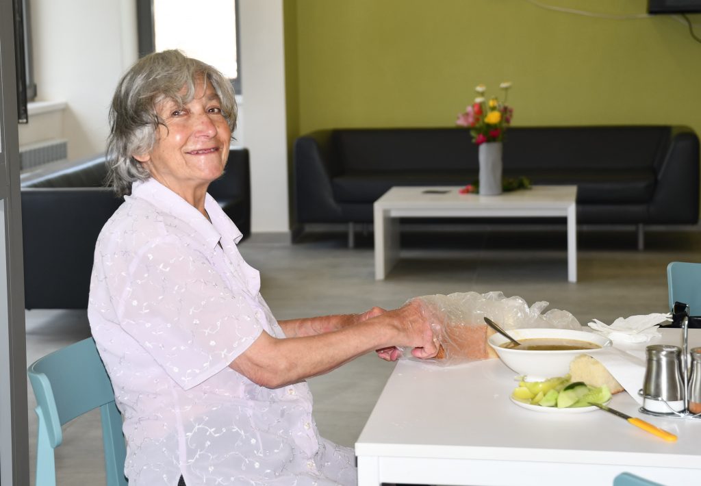 A smiling elderly woman sits at a table; on the table are a bowl of soup, some bread and some vegetables