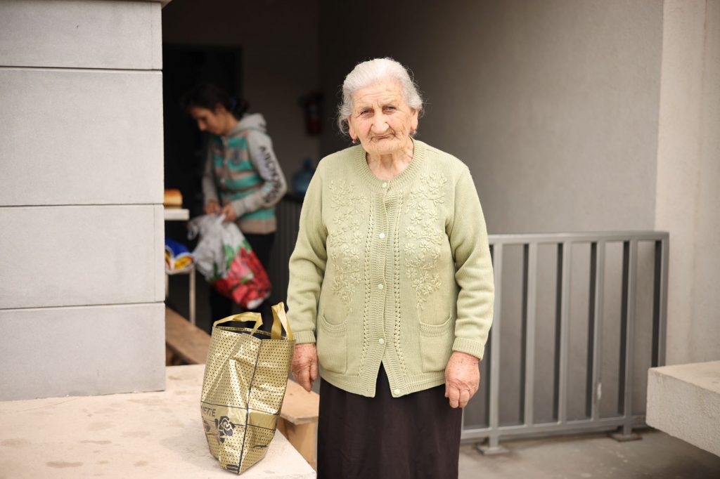 An elderly woman stands in front of a building entrance. Next to her stands a shopping bag.